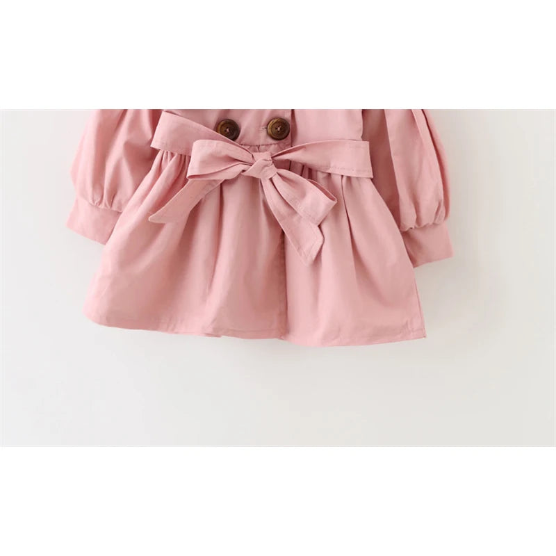 closeup på bottendelen av en rosa trench coat kappa mot en vit bakgrund. Kappan har en rosett bunden bälte i midjan och är stilren